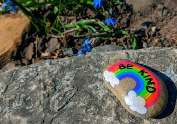 单词是善良的，彩虹涂在卵石上，背景上有春天的蓝色花朵。