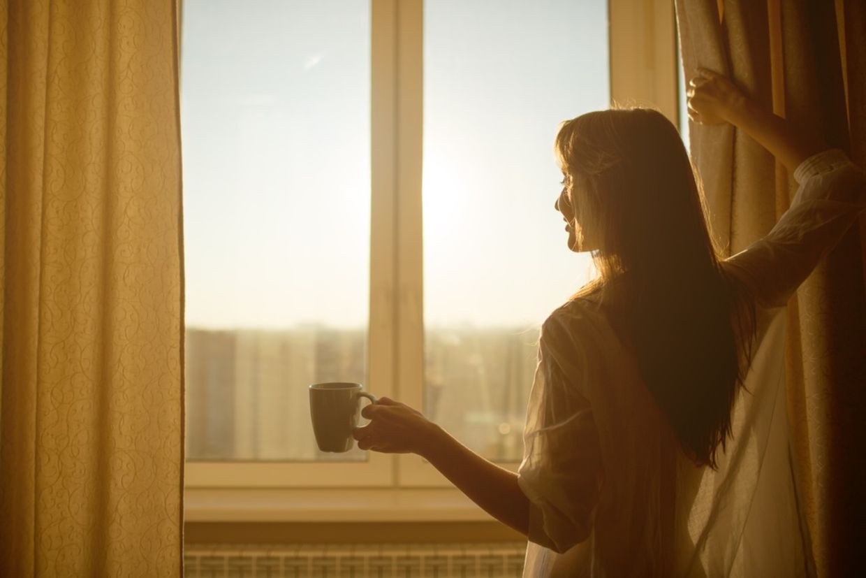 女人早上。有整洁的身体的可爱的性感的妇女拿着一个杯子用热的茶或咖啡和看的日出站立在窗口附近的在她的家和有完善的舒适早晨。