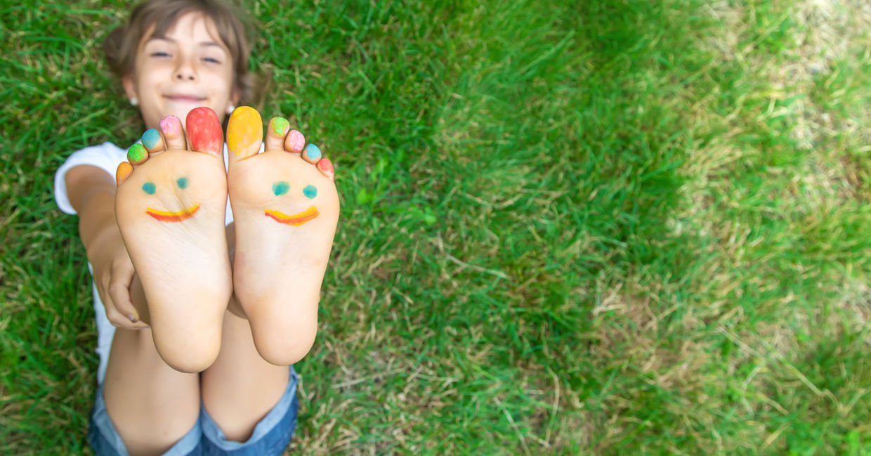 一个孩子躺在草地上，脚朝上，涂着颜料的脚趾，脚上有一张幸福的脸。