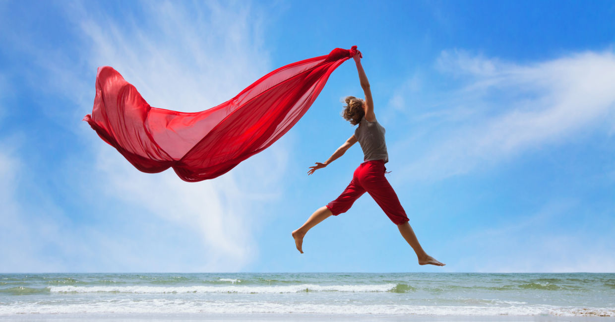 一个快乐，充实的妇女跳在海滩上，落在空中亮起一个明亮的红色围巾。